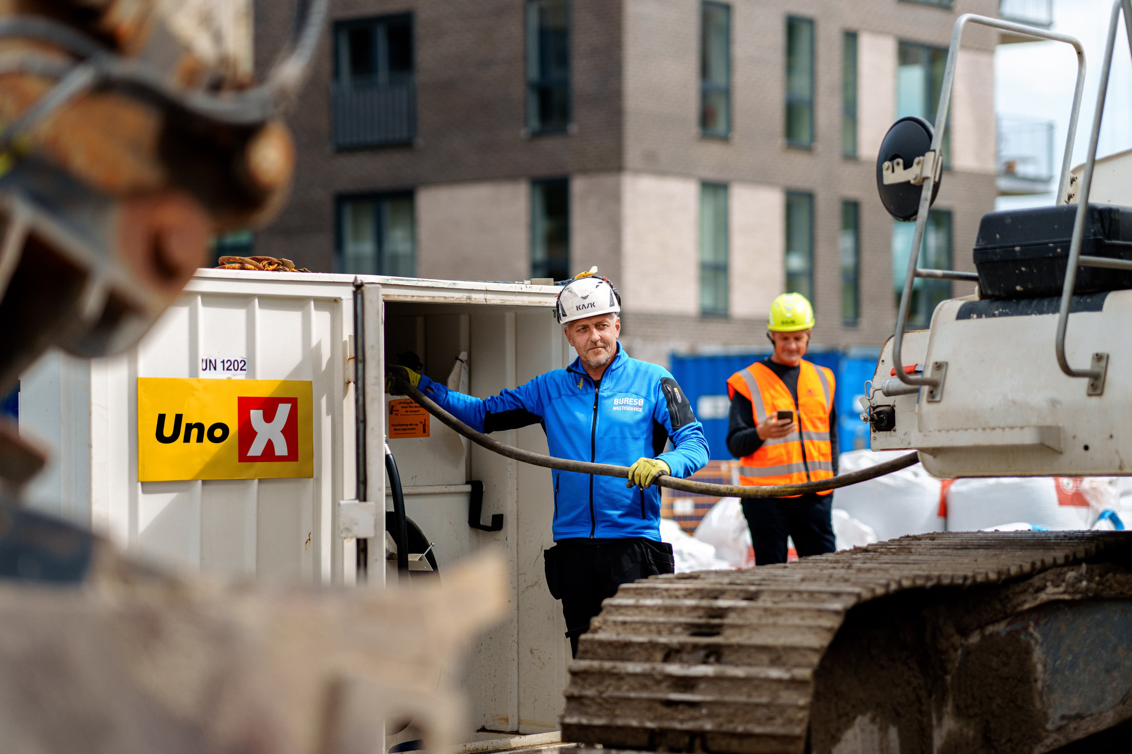 Arbejder i blå jakke fylder diesel på maskiner fra Uno-X tankcontainer, med kollega i orange refleksvest i baggrunden på byggeplads.