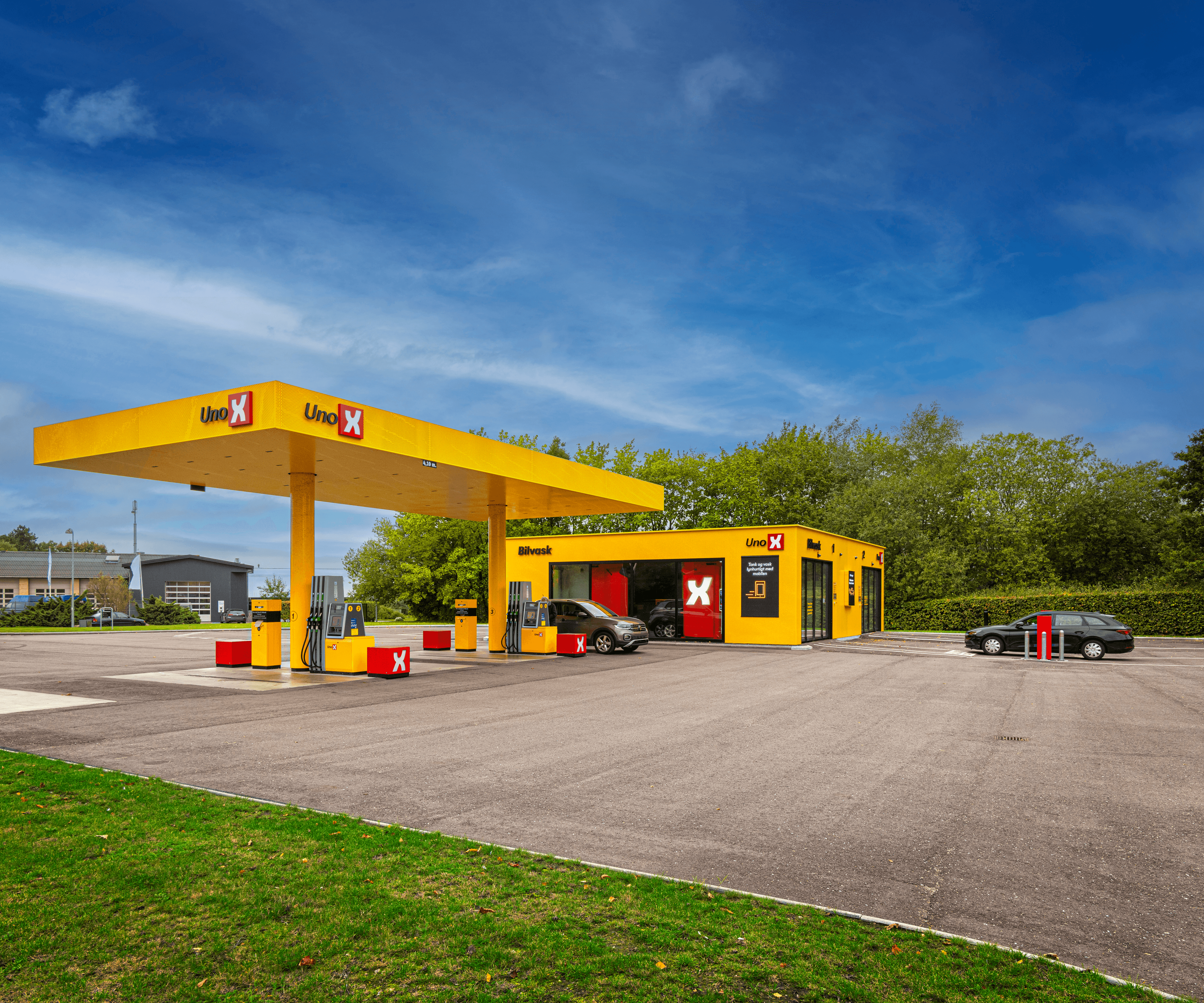 Et gult Uno X-anlæg med både tankstation og bilvask under en klar blå himmel. På venstre side ses bygningen med glasfacader og teksten "Bilvask," mens højre side viser benzinstationens standere og det overdækkede område